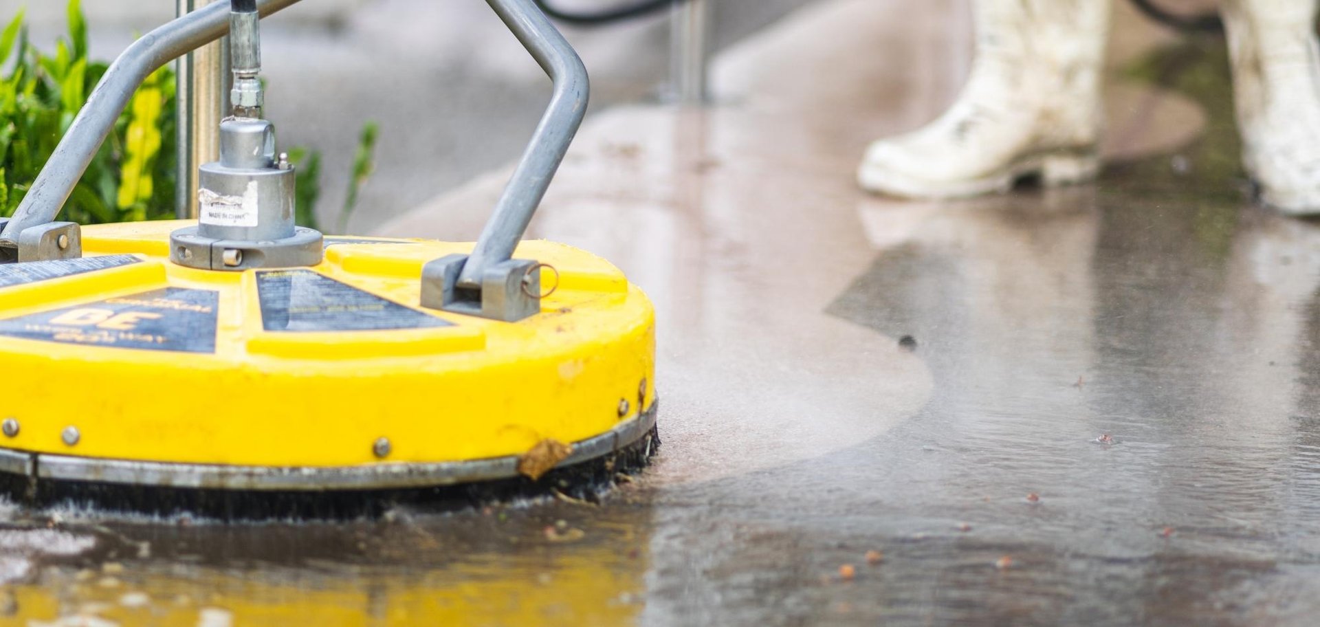 Driveway Cleaning 1 AustClean professional driveway cleaning with yellow industrial surface cleaner – specialist equipment spraying water on wet concrete pavement with technician in white boots, showcasing commercial-grade cleaning for Australian properties.