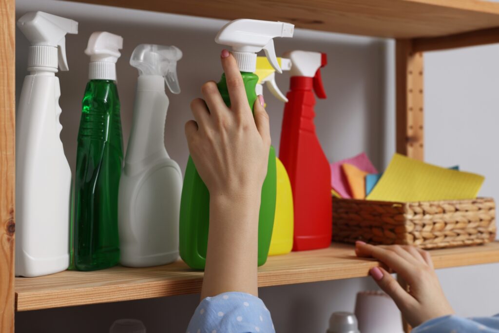 Hand reaching for green spray bottle from wooden shelf display of colorful cleaning products and cloth basket