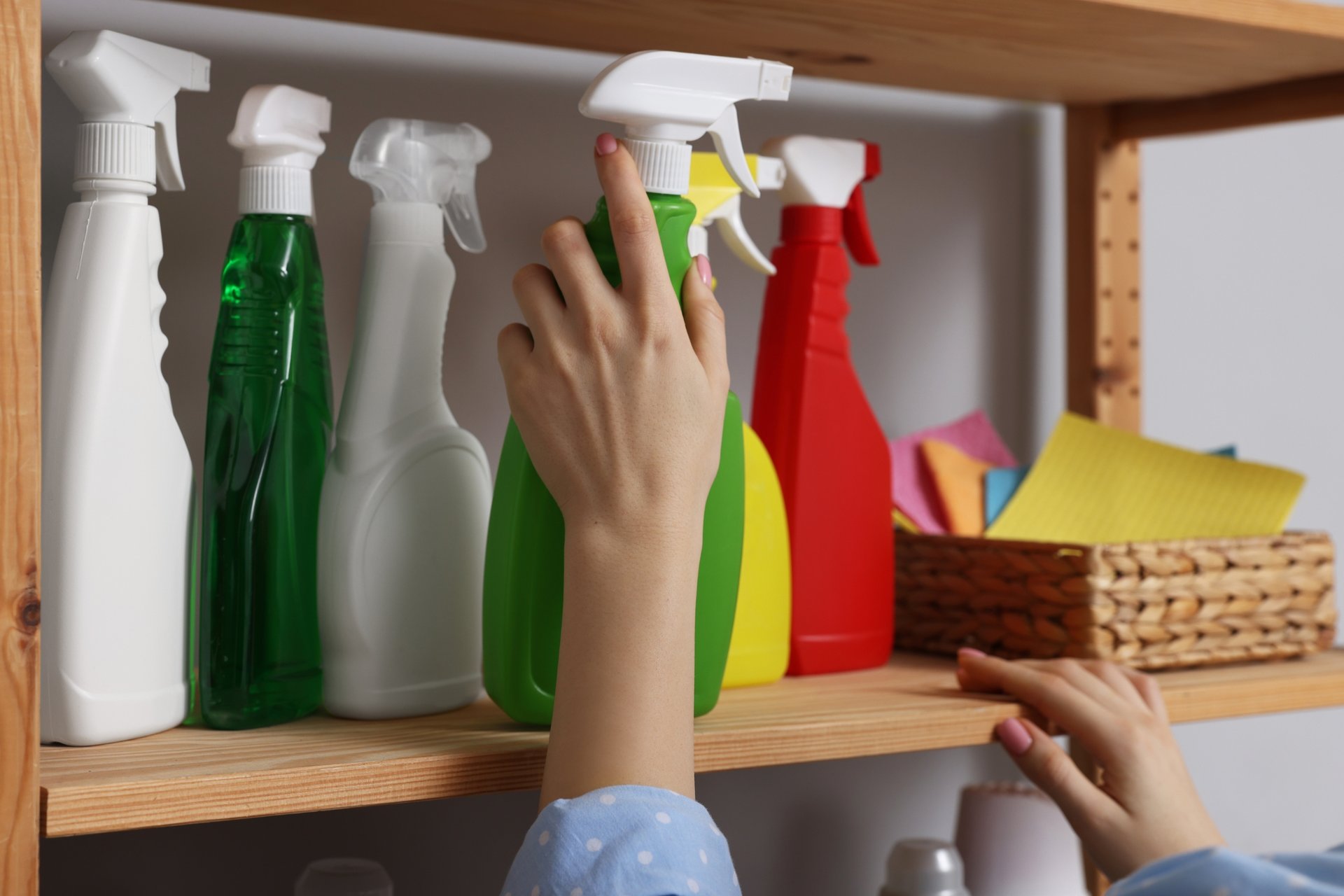 The True Cost of DIY Cleaning: Are You Really Saving Money? 1 Hand reaching for green spray bottle from wooden shelf display of colorful cleaning products and cloth basket