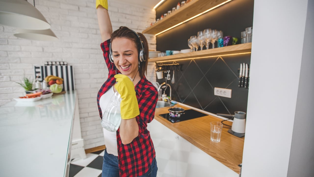 How often should you book a home cleaning service? 2 A woman wearing yellow cleaning gloves and headphones dances joyfully in a modern kitchen while holding a spray bottle, showing that even simple cleaning tips can make chores more enjoyable, with shelves of glassware and kitchen utensils visible in the background.