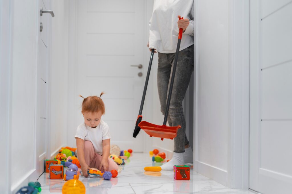 Toddler playing with colourful toys on hallway floor while adult holds broom ready for child-safe house cleaning Mountain Creek