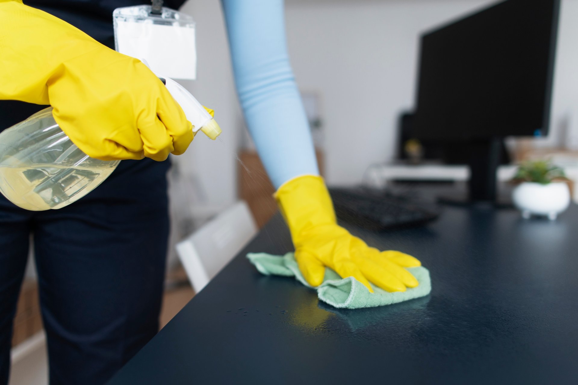 Top 3 Benefits of Office Cleaning for Merrimac Businesses 1 Person in yellow gloves applying green cloth to dark desk surface near computer monitor and keyboard workspace sanitization