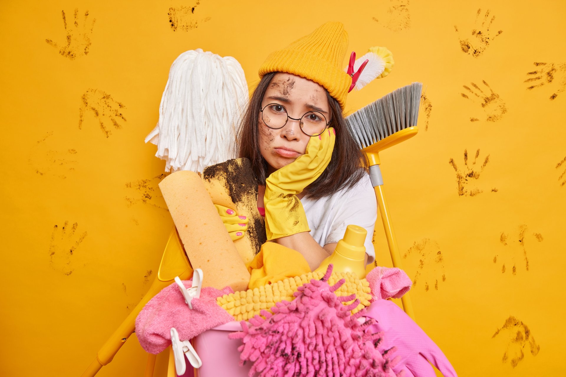 The Ultimate Australian Spring Cleaning Checklist: A Room-by-Room Guide 1 Overwhelmed woman in yellow gloves and cap holding cleaning products against muddy handprint wall expressing cleaning frustration