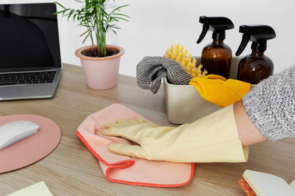 Yellow-gloved hand wiping wooden desk surface with pink cloth beside laptop and cleaning caddy